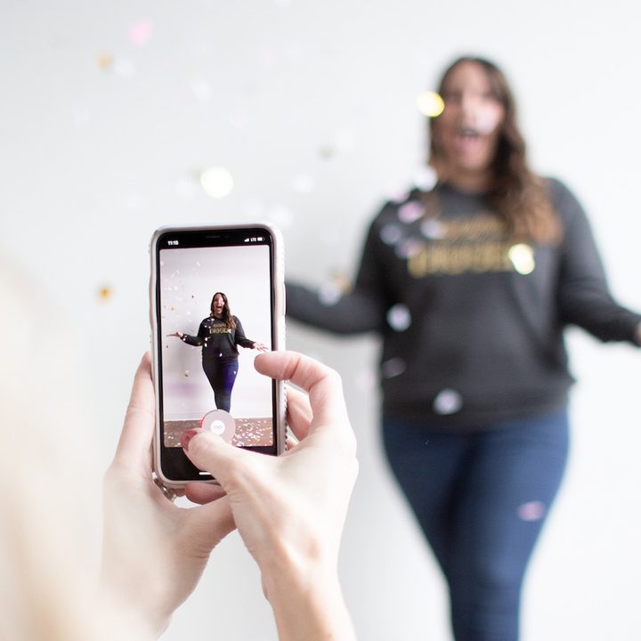 Una persona sostiene con sus manos un teléfono iPhone mientras fotografía a una mujer. Conoce cómo usar el modo retrato en iPhone