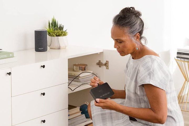 Una mujer sentada frente a un mueble blanco con la puerta abierta y conectando un cable a dispositivo Echo mientras aprende cómo configurar Alexa