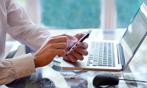Un hombre sosteniendo un celular en sus manos frente a una laptop para aprender cómo transferir tus archivos desde Android a PC