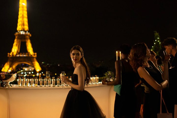 Una chica con un vestido negro, una copa en su mano y tiene en la parte trasera la torre de Paris