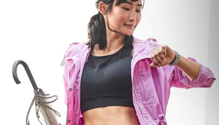Una mujer con ropa de ejercicio, un paraguas en la mano y viendo su reloj inteligente, para comparar Fitbit Versa 3 vs. Fitbit Versa 2