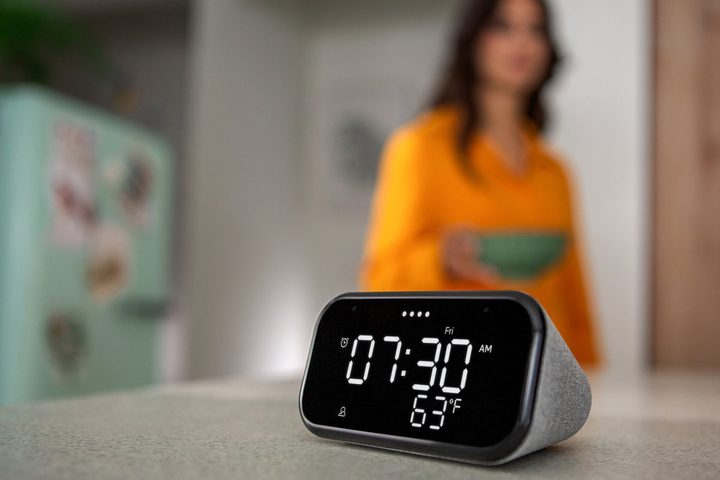 Lenovo Smart Clock Essential sobre una mesa y al fondo una mujer vestida de naranja