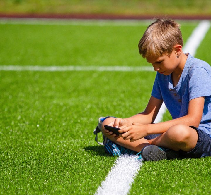 La imagen muestra a un niño sentado en una cancha mientras mira su teléfono celular.