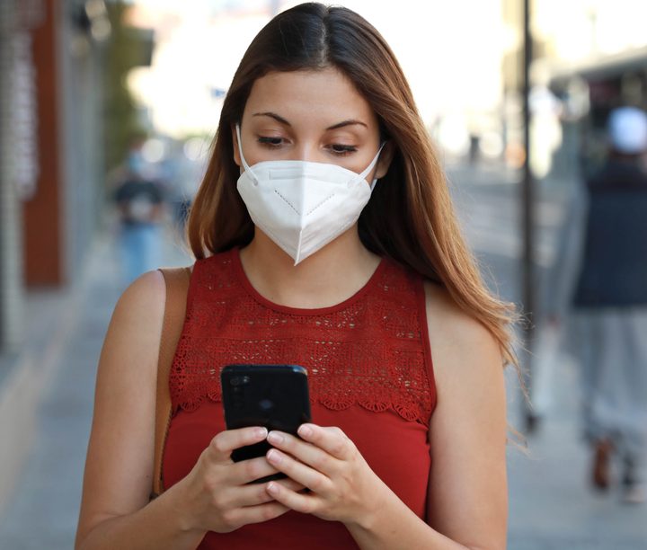 La imagen muestra a una joven con mascarilla revisando su celular.