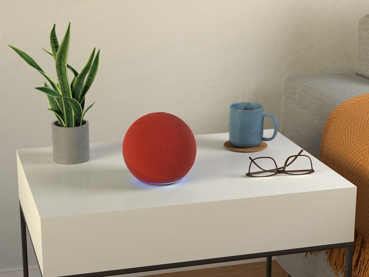 Altavoz Amazon Echo de color rojo sobre una mesita de noche junto a unas gafas, una taza y una planta