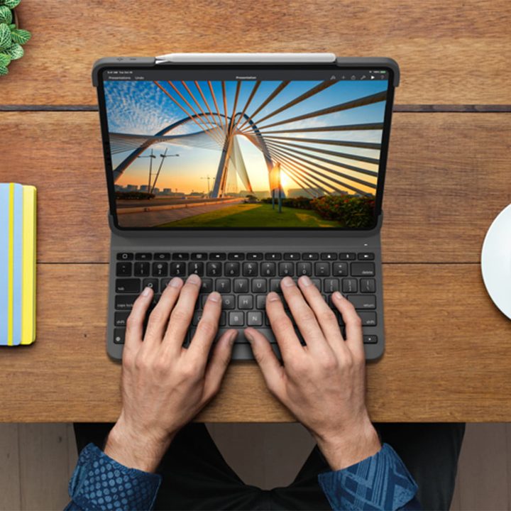 Las manos de una persona escribiendo en el Logitech Slim Folio Pro sobre una mesa a un lado de una taza y una libreta de notas, el mejor estuche iPad Pro