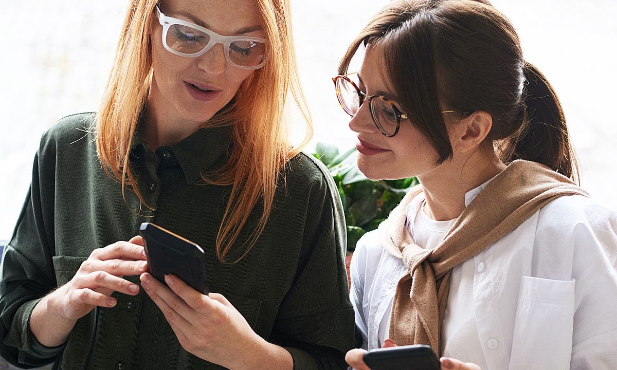 Una mujer con un teléfono con Android 12 en mano mostrándole algunos trucos a otra mujer.