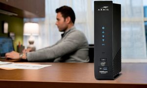 Combo de router y módem Arris Surfboard sobre un escritorio de madera y al fondo un hombre trabajando en su computadora.