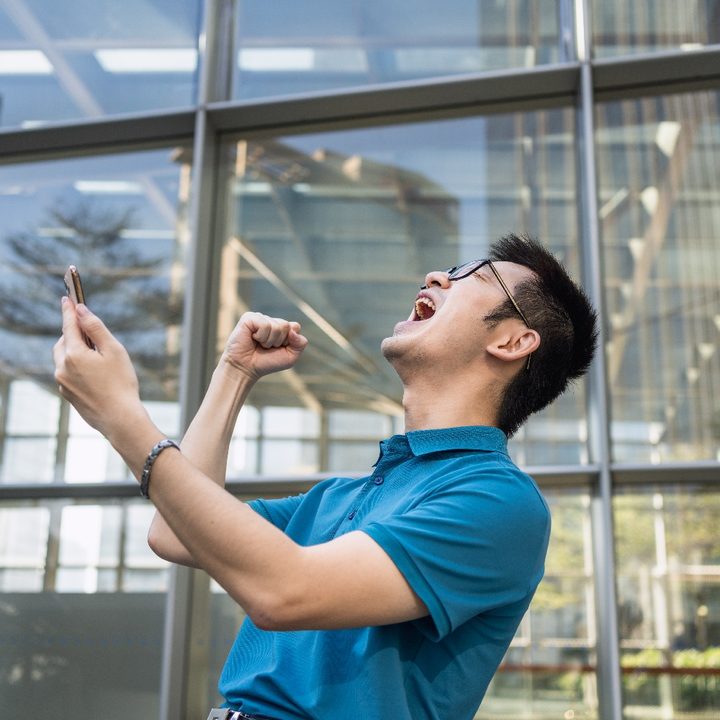 Un asiático sostiene su teléfono mientras hace un gesto de triunfo en las afueras de un edificio con paredes de vidrio