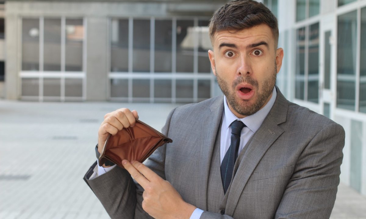 Un hombre de traje mira con preocupación mientras sostiene una billetera vacía sin dinero ni documentos.