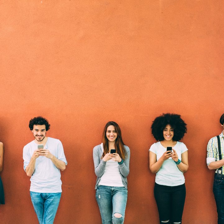 Cinco personas escribiendo mensajes de texto en sus teléfonos mientras están apoyados en una pared de color rojo