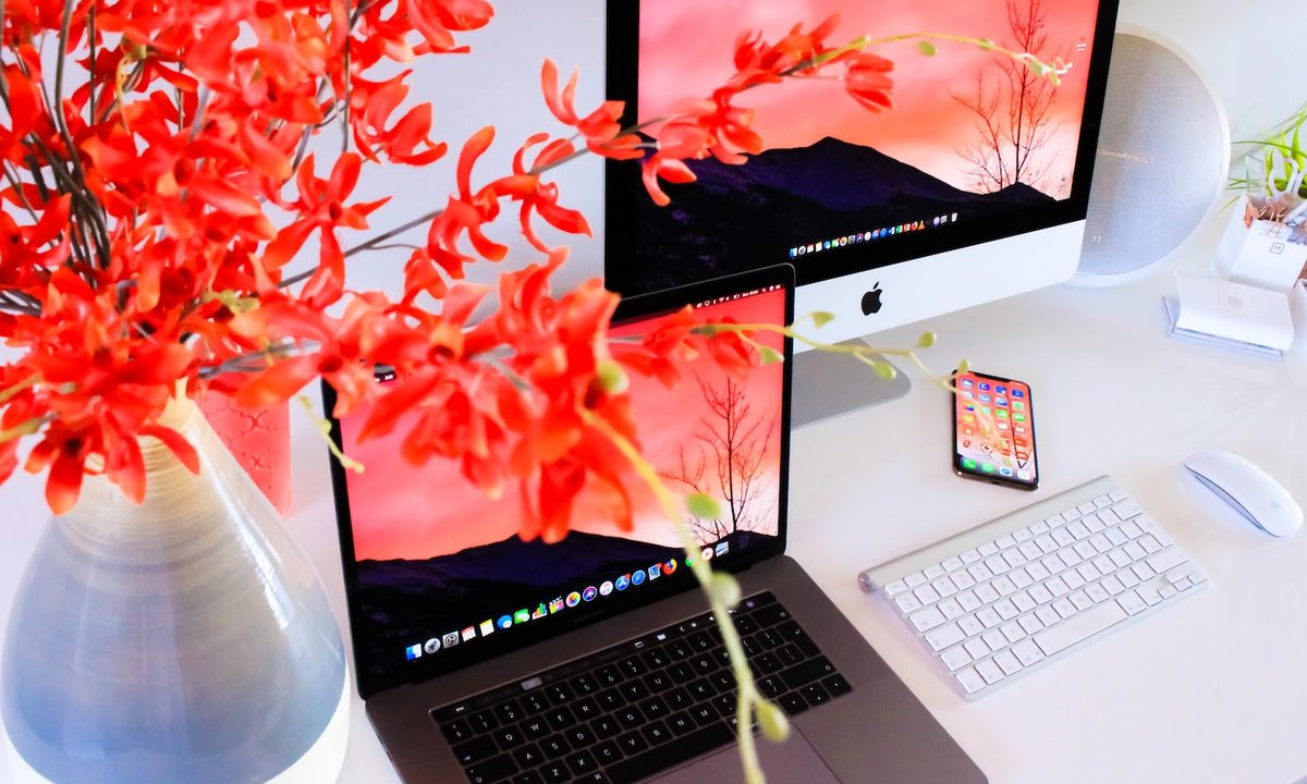 Un Macbook, un iMac , un iPhone y un teclado inalámbrico de Apple junto a un florero de colores vivos.
