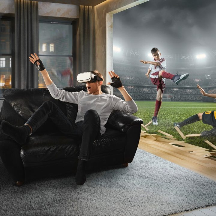 Un hombre sorprendido mientras usa visores de realidad virtual en la sala de estar de su casa, mientras en la pantalla se proyecta un partido de fútbol.