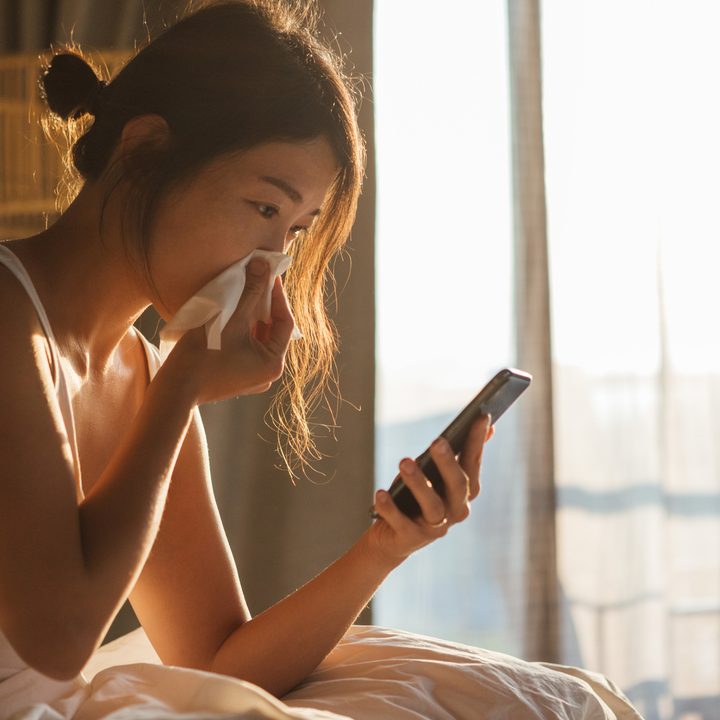 La imagen muestra a una mujer con un pañuelo en su nariz mientras revisa su celular.