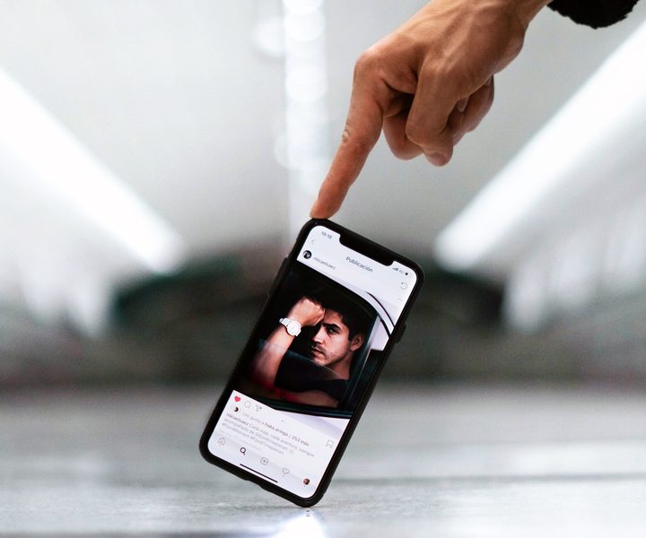 Un teléfono es sostenido con un dedo en el suelo de un salón grande e iluminado. En la pantalla se ve Instagram.