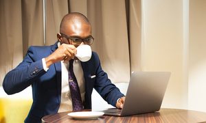 Un hpmbre tomando una taza de café mientras trabaja en su laptop.