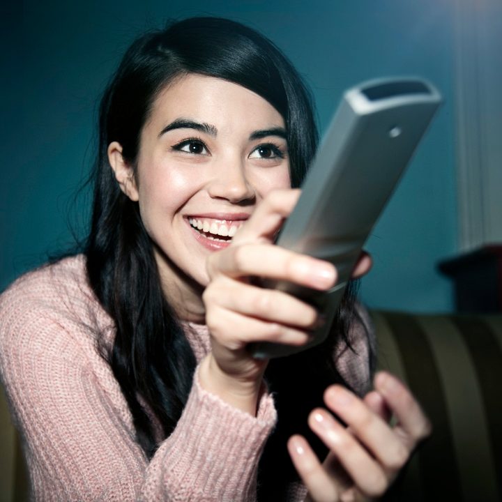 Una mujer sonríe mientras mira sostiene un control remoto y mira televisión