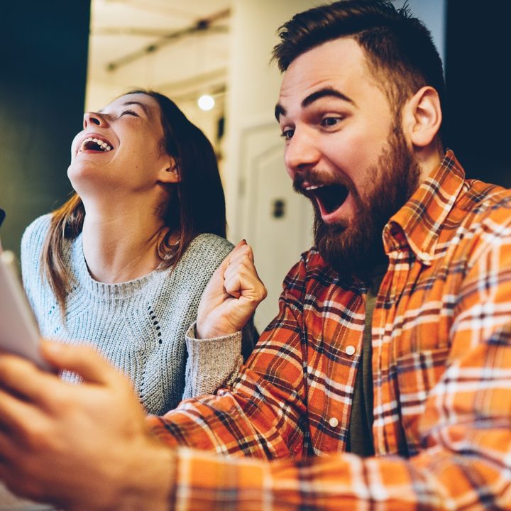 Un hombre con gafas mira sorprendido su teléfono, mientras la mujer que está a su lado derecho sonríe y celebra.