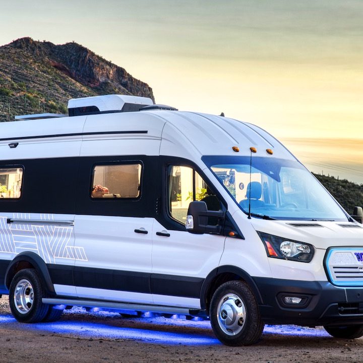 La imagen muestra a la Winnebago e-RV en color blanco estacionada con fondo de montañas y un cielo en atardecer.