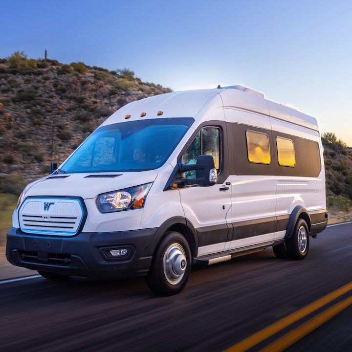 La imagen muestra a la Winnebago e-RV en color blanco circulando por una carretera con fondo de montañas.