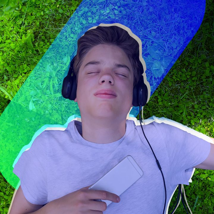 Un joven acostado sobre el césped con unos audífonos con cable y su celular.