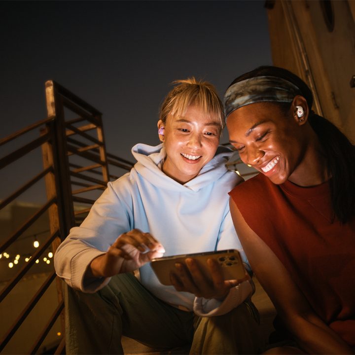 Un par de mujeres mirando el celular y con unos audífonos Beats Fit Pro.
