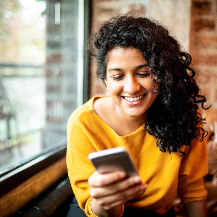 Una mujer sentada en una cafetería sonríe mientras mira la pantalla de su teléfono.