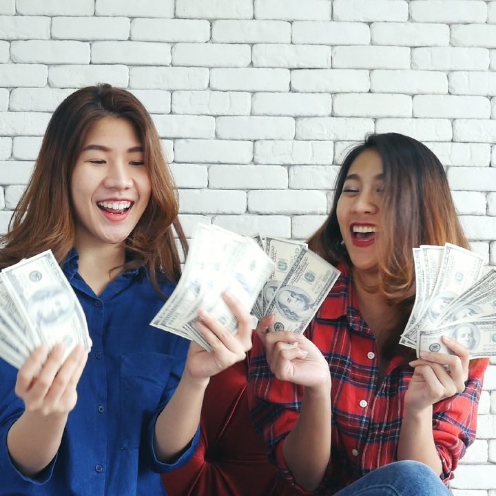 Dos mujeres sonríen mientras sostienen fajos de billetes en una sala de estar