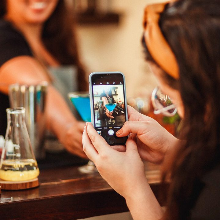 Una mujer toma una foto con su celular de un trago azul en una copa sostenida por la persona que lo preparó.