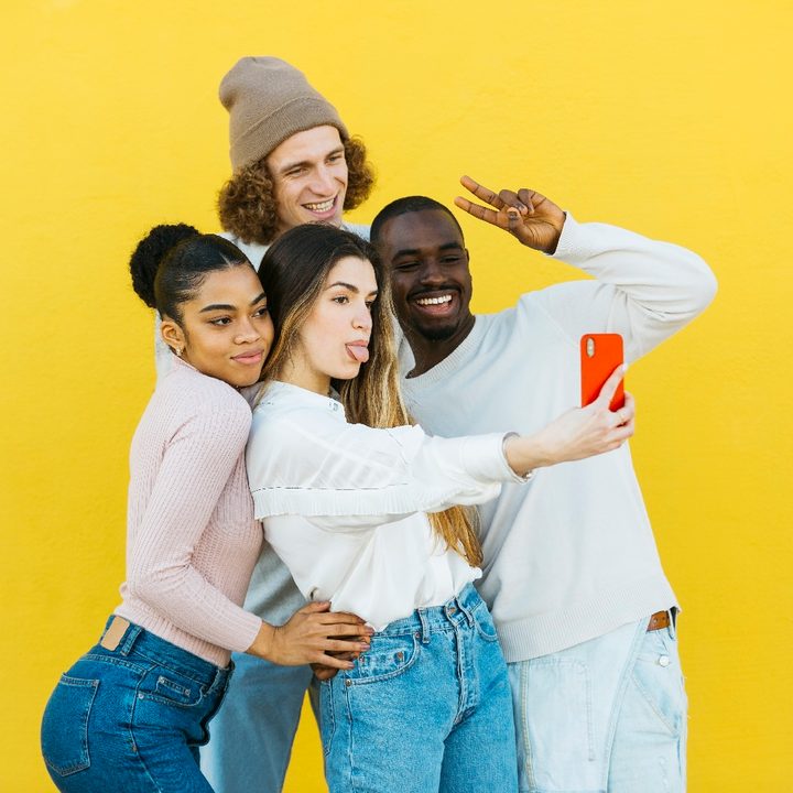 Dos mujeres y dos hombres jóvenes se toman una fotografía grupal con un teléfono inteligente con una pared amarilla de fondo.
