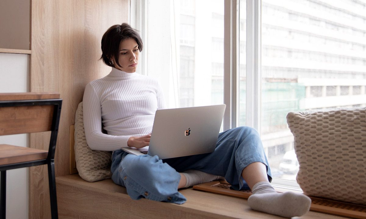 Una mujer trabaja concentrada en una laptop MacBook sentada junto a una ventana.