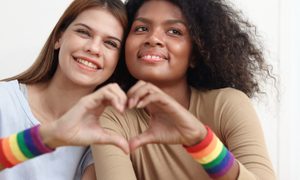 Dos chicas con pulseras LGBT haciedno un corazon con sus dedos – Las apps LGBT de citas más populares en iOS y Android.