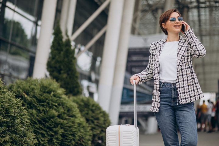 Cosas que debes hacer en tu teléfono antes de viajar al extranjero. Mujer con una maleta y hablando por teléfono afuera del aeropuerto.