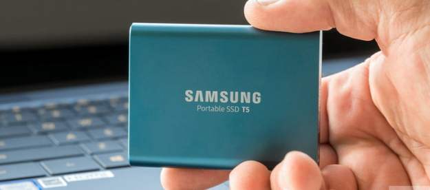 Disco duro externo Samsung T5 en la mano deunapersona y de fondo una portatil