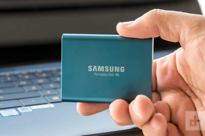 Disco duro externo Samsung T5 en la mano deunapersona y de fondo una portatil