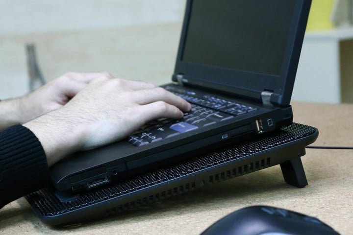 Una laptop sobre una base de enfriamiento y las manos de una persona escribiendo en ella. Si tienes un PC ruidoso, te decimos como silenciarlo