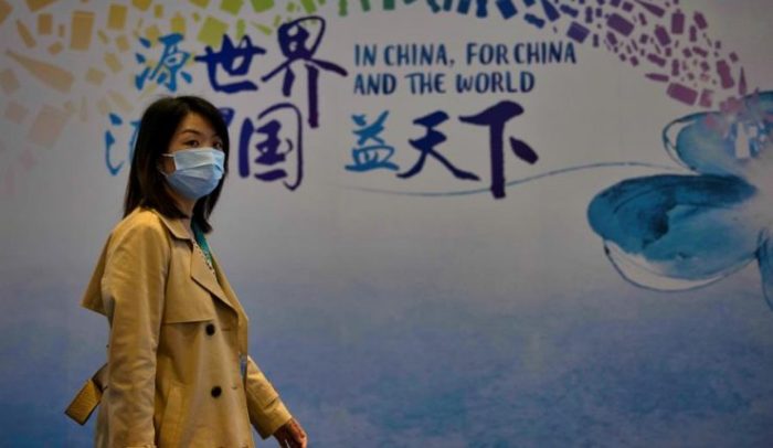 La imagen muestra a una mujer china caminando con una mascarilla en su rostro.