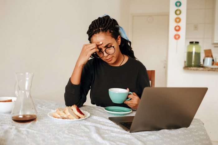 Una mujer de expresión pensativa sistiene una taza de café sentada frente a una laptop en su cocina.