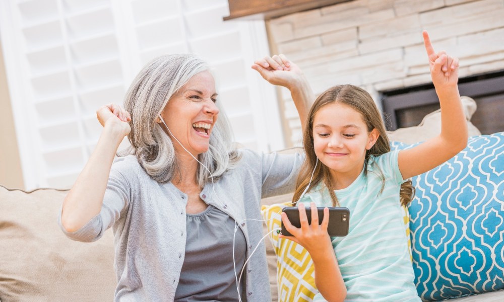 Una abuela y su nieta ríen y celebran mientras usan audífonos para escuchar música desde un teléfono celular.