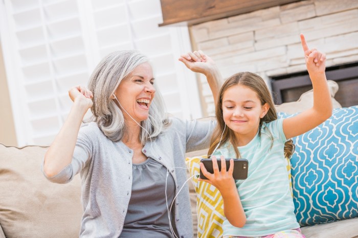 Una abuela y su nieta ríen y celebran mientras usan audífonos para escuchar música desde un teléfono celular.
