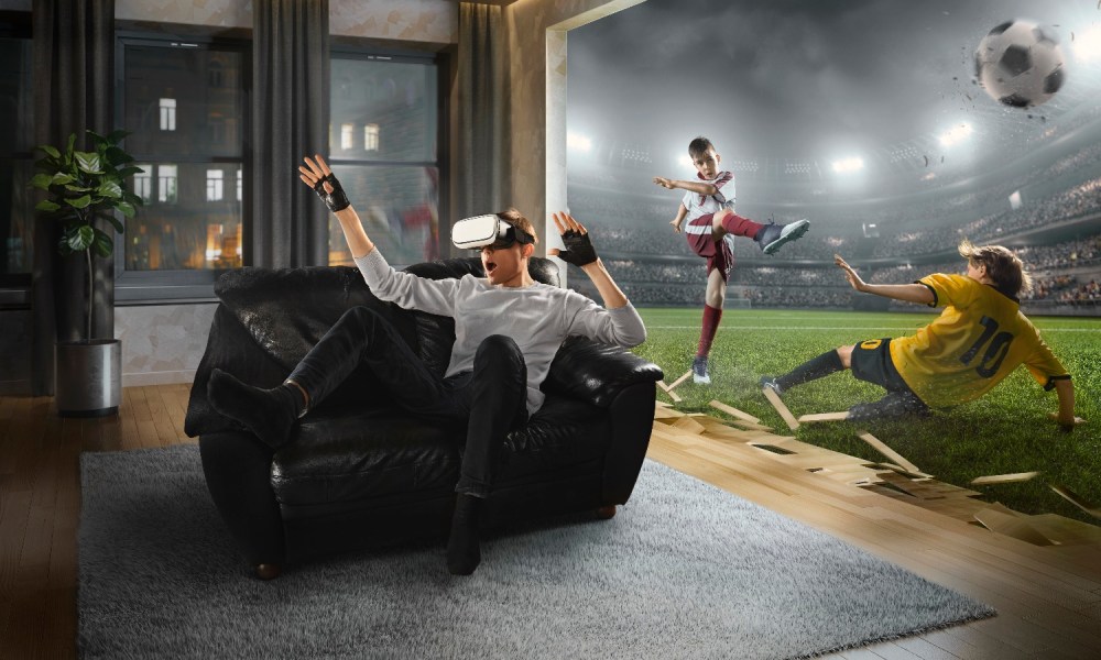 Un hombre sorprendido mientras usa visores de realidad virtual en la sala de estar de su casa, mientras en la pantalla se proyecta un partido de fútbol.