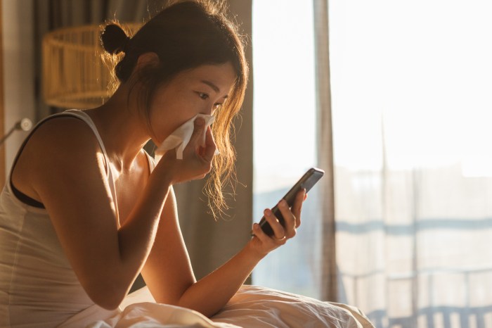 La imagen muestra a una mujer con un pañuelo en su nariz mientras revisa su celular.