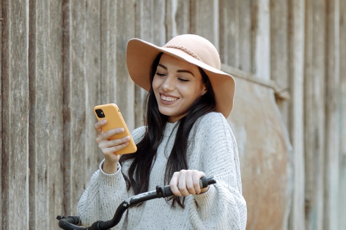 Una chica con un sombrero mira felizmente un iPhone amarillo en su mano. En la otra mano sostiene el manubrio de una bicicleta.