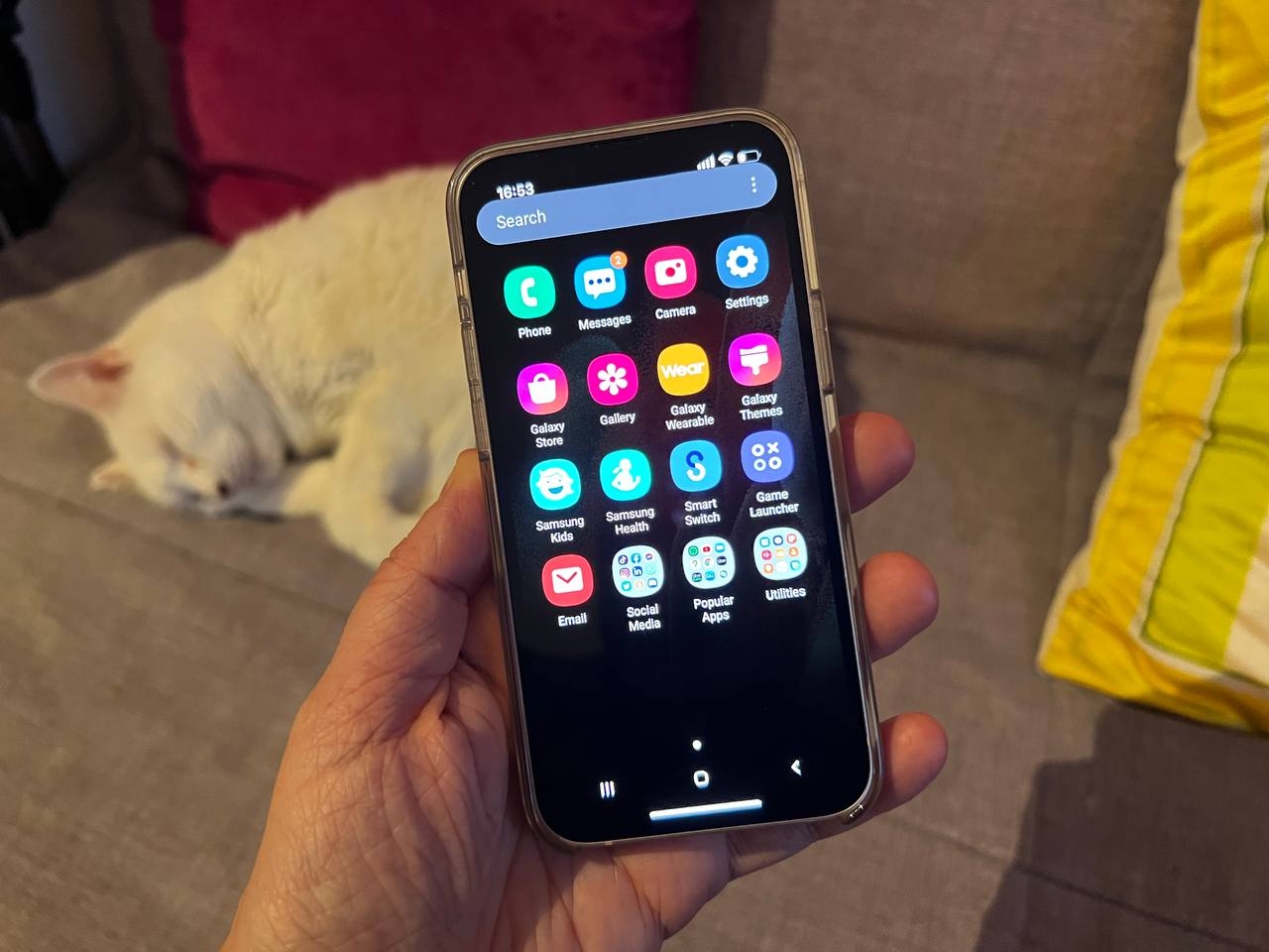 Un iPhone 13 mini sostenido con una mano y una gata blanca durmiendo de fondo. En él se puede ver el sistema operativo de Samsung.
