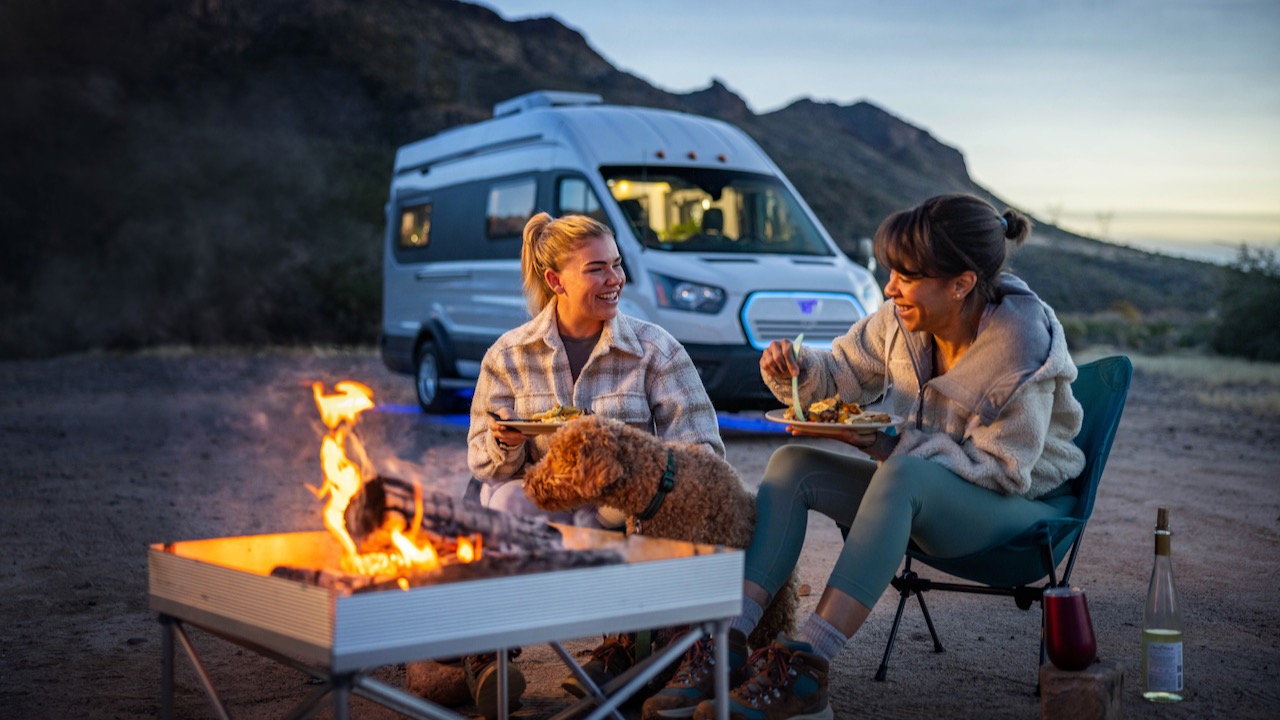 La imagen muestra a dos mujeres comiendo mientras acampan con un perro con un fondo que muestra la Winnebago e-RV en color blanc