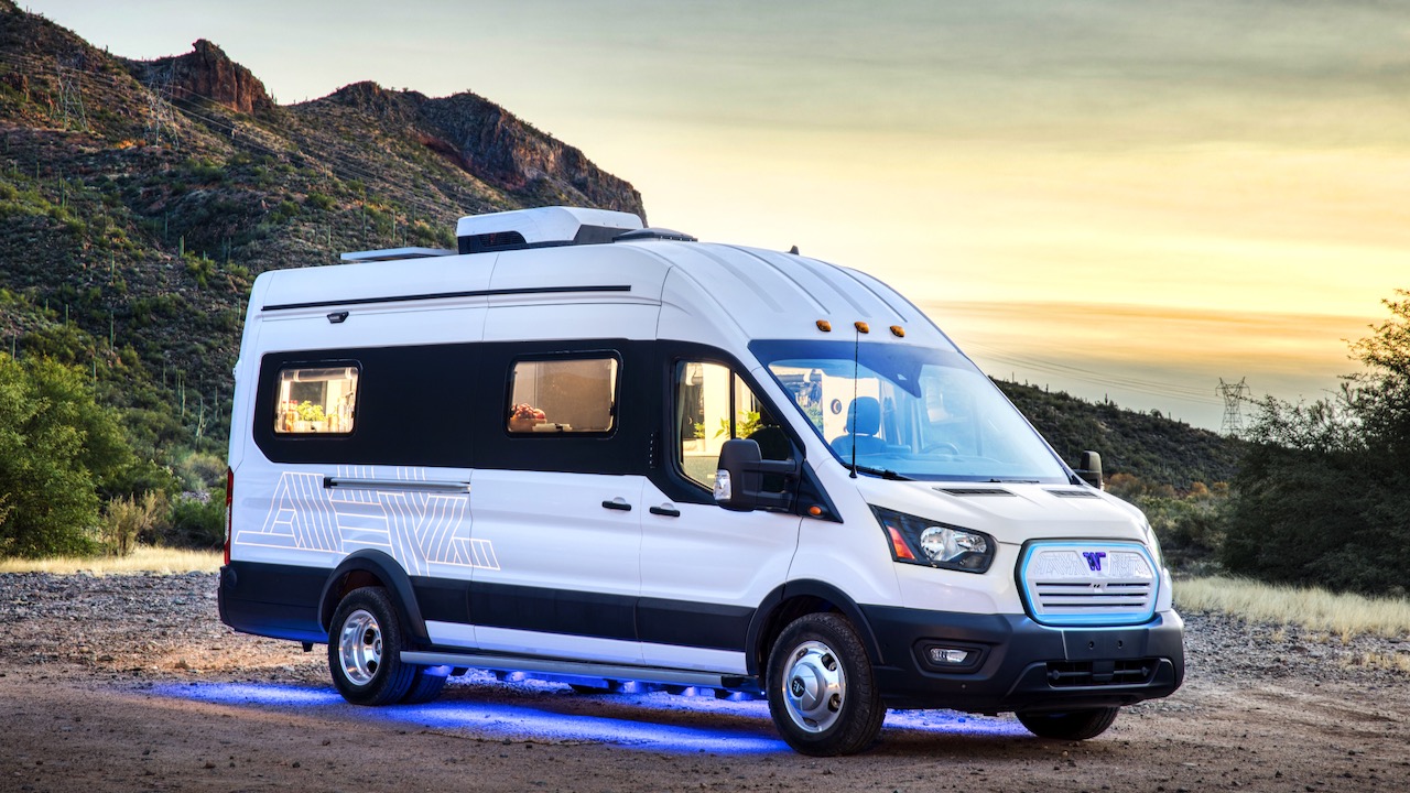 La imagen muestra a la Winnebago e-RV en color blanco estacionada con fondo de montañas y un cielo en atardecer.