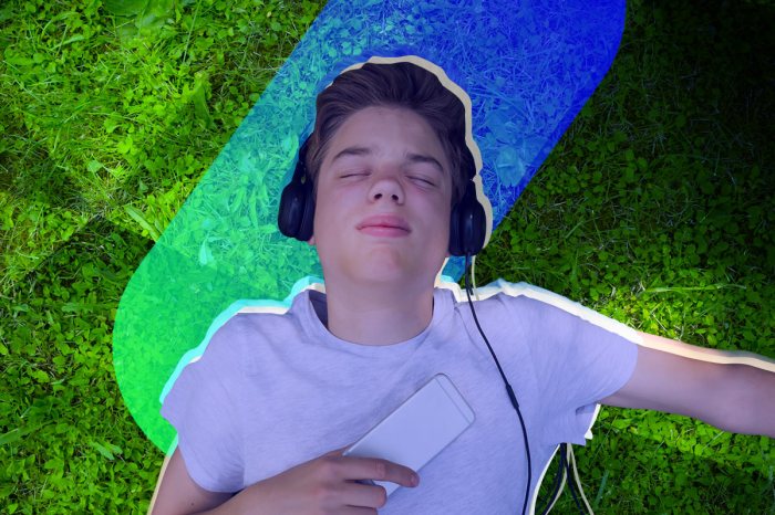 Un joven acostado sobre el césped con unos audífonos con cable y su celular.