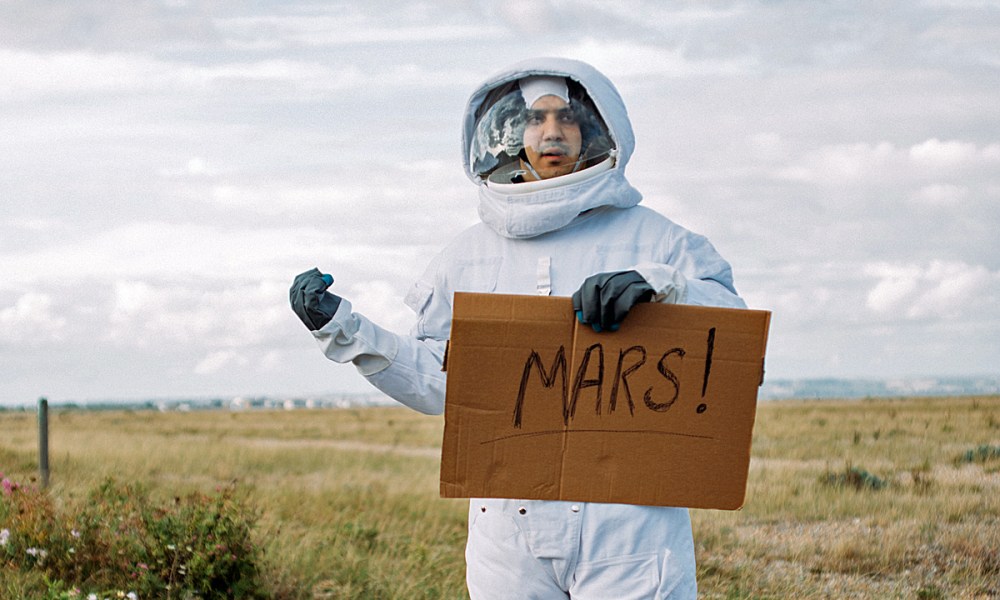 Un hombre vestido de astronauta hace autoestop con un letrero que dice MARTE.