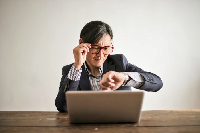 Un hombre sentado frente a una laptop observa su reloj con expresión de confusión.