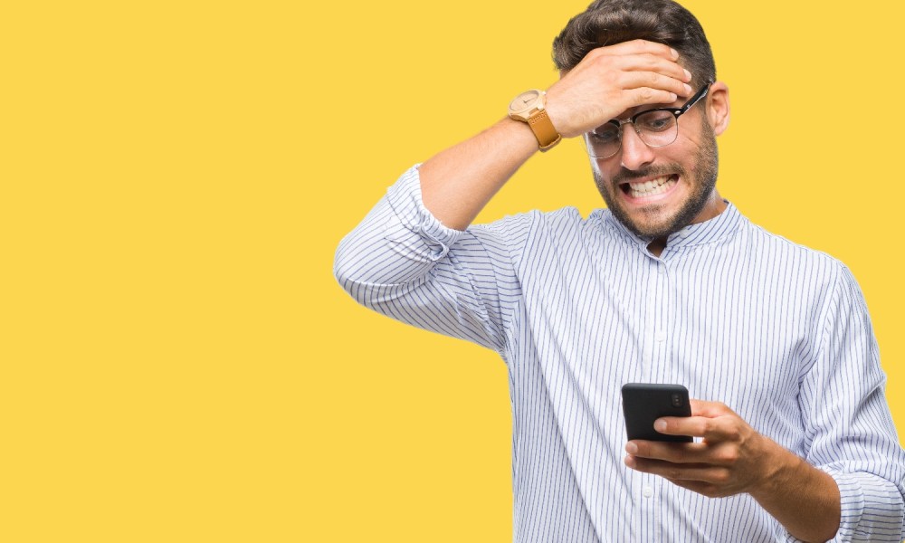 Un hombre mira con cara de preocupación su teléfono con una pared amarilla en el fondo.